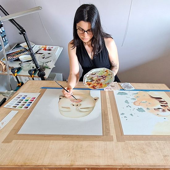 Julie Vigneron peignant à l’aquarelle dans son atelier, concentrée sur deux œuvres de la série Élément